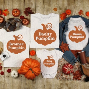 Pode incluir: T-shirts brancas e um body com gráficos de abóboras laranja e o texto "Brother Pumpkin", "Daddy Pumpkin", "Mama Pumpkin" e "Baby Pumpkin". Os artigos estão dispostos sobre uma superfície de madeira com decorações de outono.