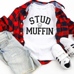 May include: White t-shirt with black text that reads "Stud Muffin" with a small cupcake graphic above the word "Muffin". The shirt is laid out on a white surface with a red and black plaid shirt, blue denim shorts, and white sneakers with black stripes.