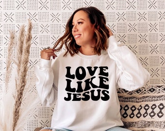 Può includere: Felpa bianca con testo nero che recita "LOVE LIKE JESUS" in un font retrò.