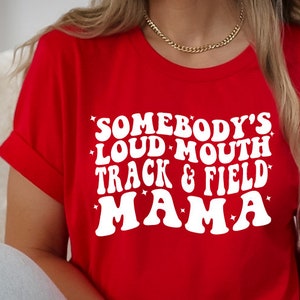 May include: Red t-shirt with white text that reads "Somebody's Loud Mouth Track & Field Mama" with stars around the text.