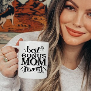 May include: White ceramic mug with black text that reads "best bonus mom ever!" in a decorative font. The mug is being held by a person.