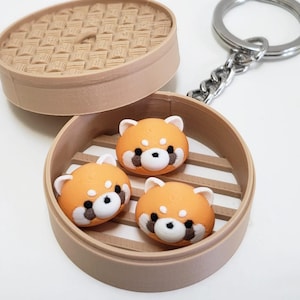 May include: A keychain featuring a miniature dim sum steamer with three orange and white red panda dumplings inside. The dumplings have black and brown facial details. The steamer is light brown and has a lid.