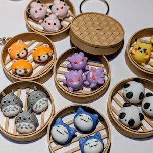 Puede incluir: Una colección de llaveros en miniatura con diferentes personajes, incluyendo pandas, Pikachu y otros personajes de dibujos animados. Los llaveros están dispuestos en pequeños recipientes redondos de estilo bambú, con una cadena plateada adjunta.