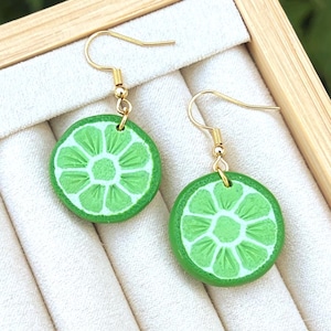 May include: A pair of lime slice earrings with gold-colored hooks. The earrings are round, with a bright green exterior and a lighter green interior design. The earrings are displayed in a wooden jewelry box.