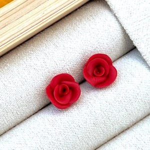 May include: A pair of small, red rose-shaped stud earrings. The earrings are displayed on a white, textured surface within a light-colored wooden frame. Yellow flowers are visible in the background.