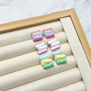 Könnte beinhalten: Eine Sammlung bunter, miniaturhafter, gestapelter Buch-förmiger Anhänger in Pastelltönen von Rosa, Lila, Blau und Grün. Die Anhänger sind auf einem beigefarbenen, stoffausgekleideten Display-Tablett mit Holzrahmen angeordnet.