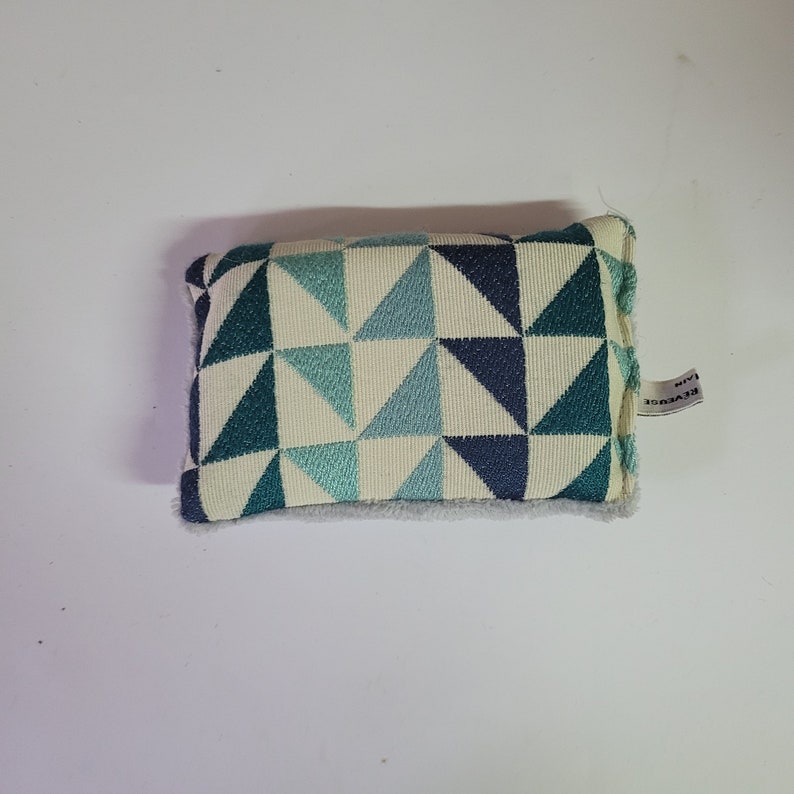May include: A small, rectangular pillow with a white background and a geometric pattern of blue and green triangles. The pillow has a white label on the side that says "Reverse" in black text.