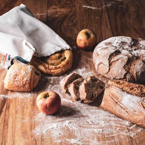 May include: A white cloth bag filled with various breads, including a round loaf, a long loaf, and a pastry. There are also two red apples on a wooden surface.