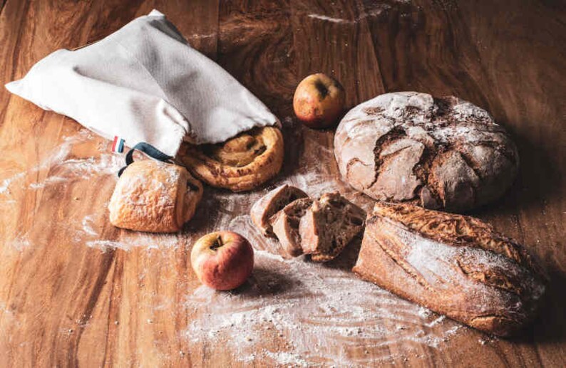 May include: A white cloth bag filled with various breads, including a round loaf, a long loaf, and a pastry. There are also two red apples on a wooden surface.
