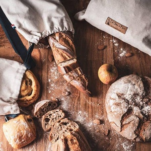 May include: A flat lay image of various breads, including a baguette, a round loaf, and a pastry, all on a wooden surface. There are also two white cloth bags with blue straps, one of which has the text "Le Sac &agrave; Pain" printed on it. The image also includes an apple and almonds.