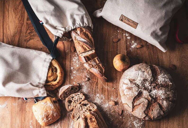 May include: A flat lay image of various breads, including a baguette, a round loaf, and a pastry, all on a wooden surface. There are also two white cloth bags with blue straps, one of which has the text "Le Sac &agrave; Pain" printed on it. The image also includes an apple and almonds.