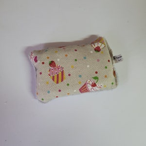 May include: A beige fabric pillow with a white border and a pattern of colorful cupcakes with pink frosting and green leaves. The pillow has a small white tag with the text "A Home Ut".