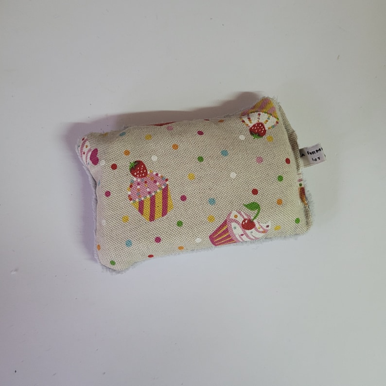 May include: A beige fabric pillow with a white border and a pattern of colorful cupcakes with pink frosting and green leaves. The pillow has a small white tag with the text "A Home Ut".