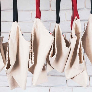 May include: Seven white canvas tote bags hanging from a white brick wall. The bags have red, white, and blue trim and are all empty.