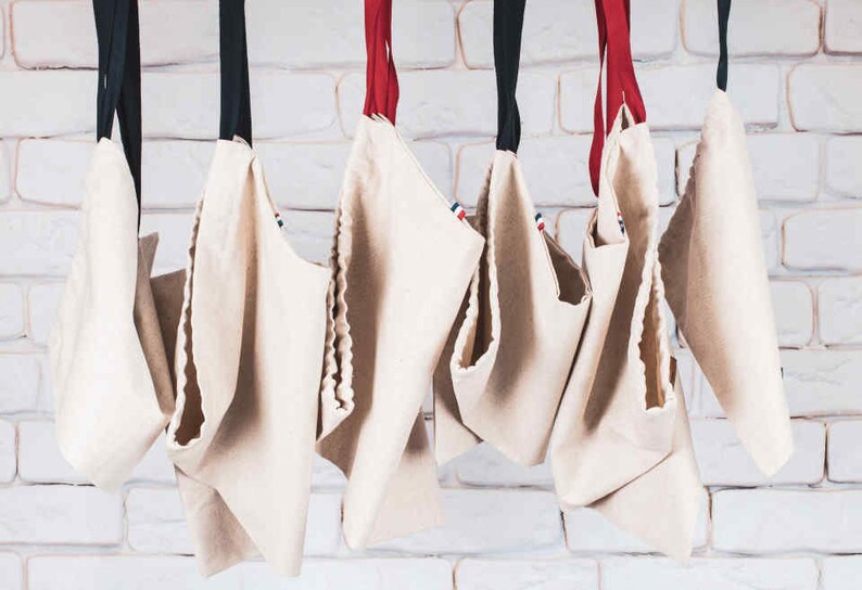May include: Seven white canvas tote bags hanging from a white brick wall. The bags have red, white, and blue trim and are all empty.