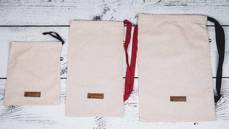 May include: Three beige drawstring bags with different sized black and red drawstrings. The bags have a brown label with the text "La Bagagerie" on them.