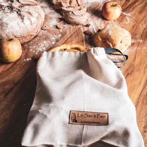 May include: A white canvas bag with a brown leather label that says "Le Sac &agrave; Pain" and "La P&acirc;tisserie Revenante". The bag is filled with bread and is sitting on a wooden table with apples and other bread.