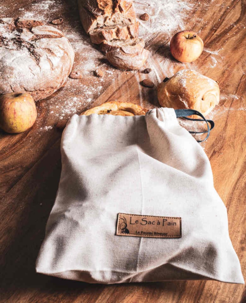 May include: A white canvas bag with a brown leather label that says "Le Sac &agrave; Pain" and "La P&acirc;tisserie Revenante". The bag is filled with bread and is sitting on a wooden table with apples and other bread.