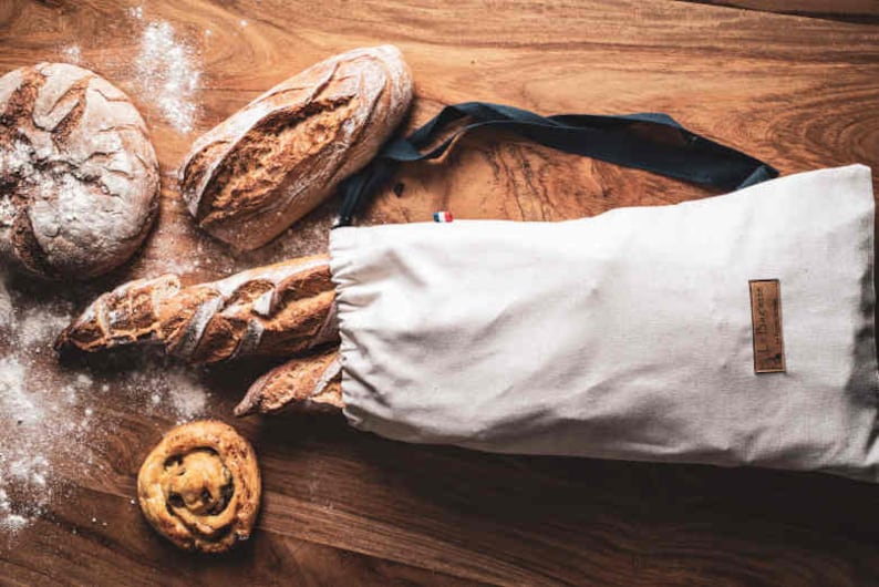 May include: A white canvas bag with a black strap and a brown label that says "Le Dunette" is filled with various loaves of bread. The bag is sitting on a wooden surface.