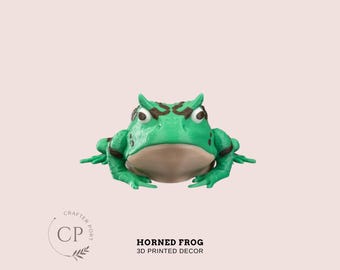May include: A 3D printed green horned frog with brown markings. The frog is sitting on a white background. The frog has a wide mouth and large eyes. The frog is a decorative item.