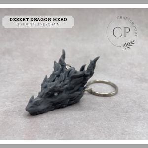 May include: A 3D printed gray dragon head keychain with a silver key ring. The keychain is labeled "Desert Dragon Head 3D Printed Keychain" and "Crafter Port CP".