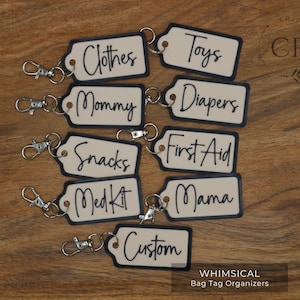 May include: Nine cream-colored bag tags with black trim and black handwritten text. The tags are labeled "Clothes", "Toys", "Mommy", "Diapers", "Snacks", "First Aid", "Med Kit", "Mama", and "Custom". Each tag has a silver key ring attached.