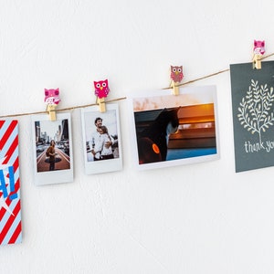Puede incluir: Una cuerda de fotos y una tarjeta est&aacute;n colgadas en una pared blanca con pinzas de ropa de b&uacute;ho de colores. Las fotos incluyen una pareja, un gato y una calle de la ciudad. La tarjeta dice "gracias".