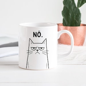 Könnte beinhalten: Weiße Keramik-Tasse mit einer schwarzen Illustration eines grimmigen Katzengesichts und dem Wort "NÖ." darüber. Die Tasse hat einen weißen Henkel. Im Hintergrund ein kupferfarbener Topf und eine grüne Pflanze.