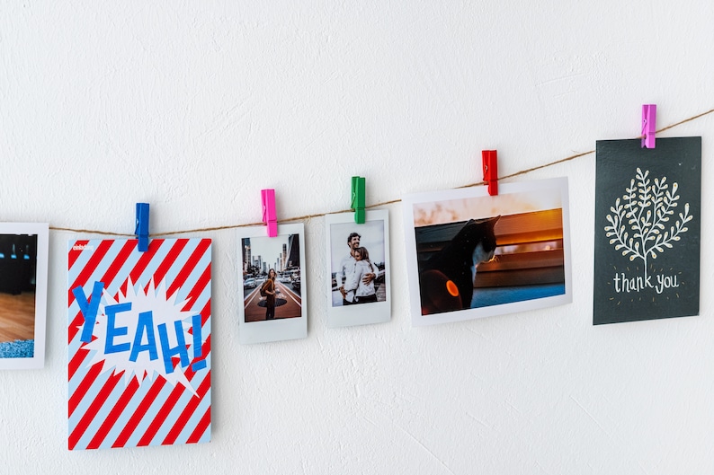 Puede incluir: Una cuerda de fotos y tarjetas cuelga de una pared blanca con pinzas de colores. Las fotos incluyen una pareja, un gato y una calle de la ciudad. Las tarjetas incluyen una tarjeta roja y blanca a rayas con la palabra "YEAH!" en azul y una tarjeta negra con un &aacute;rbol blanco y las palabras "gracias".