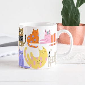 Könnte beinhalten: Weiße Keramiktasse mit bunten Cartoon-Katzen in verschiedenen Posen. Die Katzen haben unterschiedliche Muster und Farben, darunter Orange mit blauen Punkten, Rosa mit orangefarbenen Streifen und Gelb mit rosa Punkten. Die Tasse hat einen weißen Henkel.