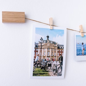 Puede incluir: Una cadena de fotos est&aacute; colgada en una pared blanca con clips de madera. Las fotos incluyen un edificio con un cielo azul, una persona de pie en una playa y una tarjeta roja y blanca a rayas con la palabra "S&iacute;" en azul.
