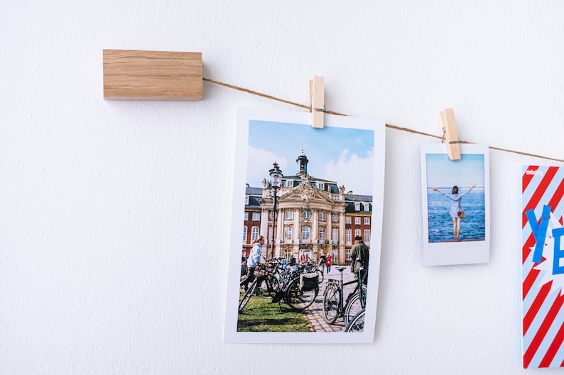 Puede incluir: Una cadena de fotos est&aacute; colgada en una pared blanca con clips de madera. Las fotos incluyen un edificio con un cielo azul, una persona de pie en una playa y una tarjeta roja y blanca a rayas con la palabra "S&iacute;" en azul.