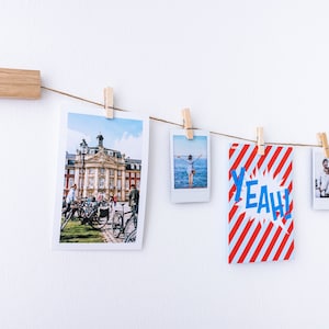 Puede incluir: Una cuerda con fotos y una tarjeta con rayas rojas y blancas y la palabra "YEAH!" en letras azules cuelgan de una pared blanca con pinzas de madera.
