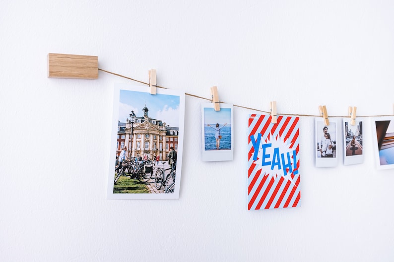 Puede incluir: Una cuerda con fotos y una tarjeta con rayas rojas y blancas y la palabra "YEAH!" en letras azules cuelgan de una pared blanca con pinzas de madera.