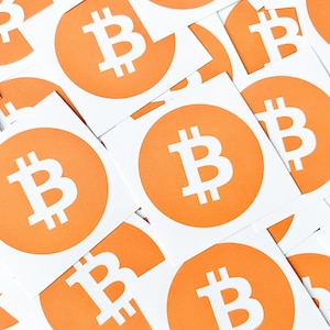 Könnte beinhalten: Nahaufnahme von mehreren weißen quadratischen Aufklebern mit orangenen Kreisen, die das Bitcoin-Symbol in Weiß enthalten.