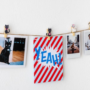 Puede incluir: Una cuerda de fotos y una tarjeta con rayas rojas y blancas y la palabra "YEAH!" en letras azules cuelgan de una pared blanca con pinzas para la ropa de gato blanco y negro.