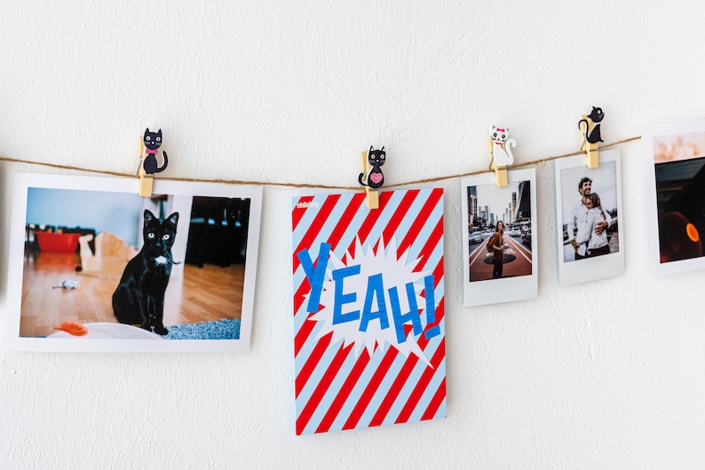 Puede incluir: Una cuerda de fotos y una tarjeta con rayas rojas y blancas y la palabra "YEAH!" en letras azules cuelgan de una pared blanca con pinzas para la ropa de gato blanco y negro.