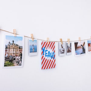 Puede incluir: Una cuerda con fotos y una tarjeta con la palabra "YEAH!" impresa en ella est&aacute;n colgadas en una pared con pinzas de madera. Las fotos son de varios temas, incluyendo una calle de la ciudad, una playa y un gato.