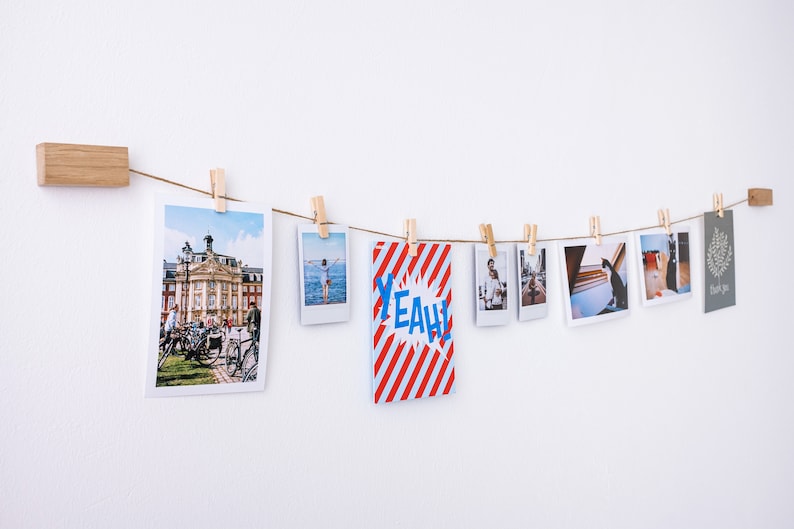 Puede incluir: Una cuerda con fotos y una tarjeta con la palabra "YEAH!" impresa en ella est&aacute;n colgadas en una pared con pinzas de madera. Las fotos son de varios temas, incluyendo una calle de la ciudad, una playa y un gato.