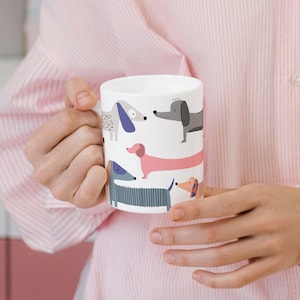 Könnte beinhalten: Weiße Keramiktasse mit einem sich wiederholenden Muster von Cartoon-Dackeln in verschiedenen Farben. Die Hunde haben lange Körper und kurze Beine. Die Tasse wird von einer Person gehalten, die ein rosa gestreiftes Hemd trägt.