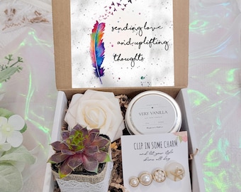 May include: A gift box filled with a succulent plant, a white rose, a small candle, and a pair of pearl hair clips. The box also includes a card with the text "sending love and uplifting thoughts".