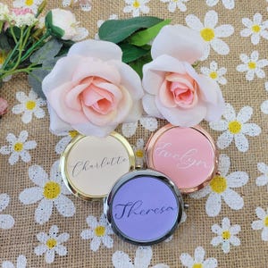 May include: Three personalized compact mirrors in gold, rose gold, and silver frames, each with a different name: "Charlotte", "Evelyn", and "Theresa". The mirrors are surrounded by pink and white roses and daisy accents.