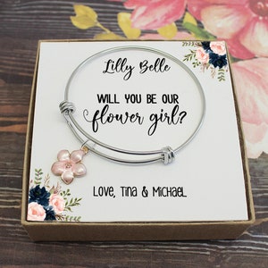 May include: A silver bracelet with a pink flower charm in a box. The box has a white card with the text "Lilly Belle, Will you be our flower girl? Love, Tina & Michael."