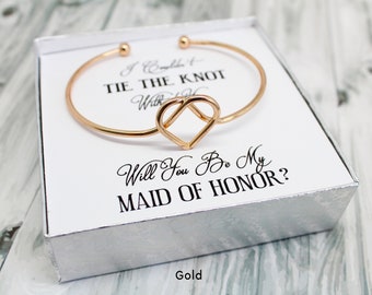 May include: Gold bracelet with a heart-shaped knot design in a white box. The box has the text "I Can't Tie The Knot Without You" and "Will You Be My Maid Of Honor?" printed on it.