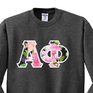 May include: A charcoal gray crewneck sweatshirt featuring the Greek letters Alpha and Phi in a white outline. The interior of the letters is filled with a vibrant floral pattern consisting of pink peonies, green leaves, and dark berries on a black background.