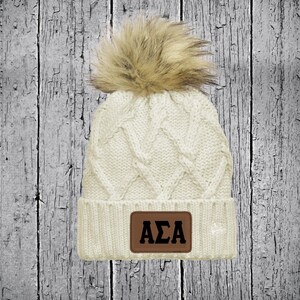 May include: White cable knit beanie with a brown faux fur pom pom and a brown leather patch with the letters "AZA" in black.