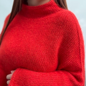 Puede incluir: Un suéter rojo vibrante con un cuello alto y acanalado. El suéter tiene mangas largas y una textura de punto grueso. El primer plano resalta la apariencia suave y cálida de la prenda, lo que sugiere un ajuste cómodo. El suéter es de color sólido.