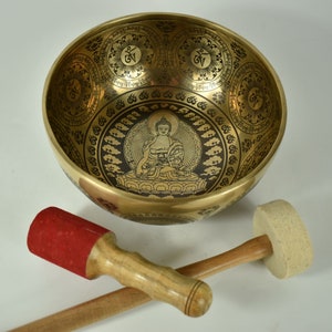 Könnte beinhalten: Eine goldfarbene Klangschale mit aufwendigen geätzten Designs, darunter eine zentrale Buddha-Figur und kreisförmige Muster. Zwei Holzschlägel mit Filzschlagflächen, einer rot und einer beige, sind enthalten.