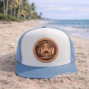 May include: A blue and white trucker hat with a leather patch showing a beach scene with palm trees and the text "Throwing Shade co." The hat is on a sandy beach with the sea in the background.