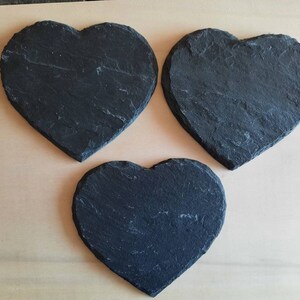 May include: Three black heart-shaped slate coasters. The coasters are made of natural slate and have a rustic, uneven texture.